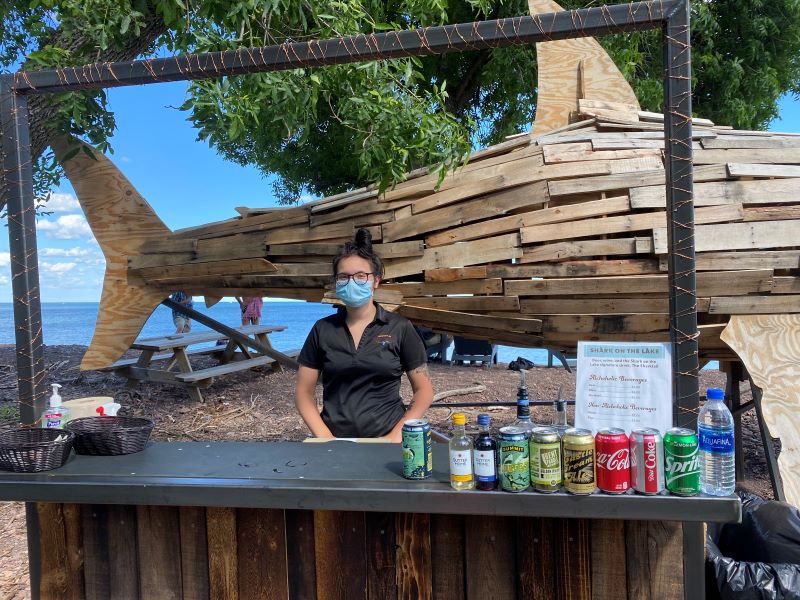 Bartender wearing a mask at an outdoor bar in front of a shark statue made of wood