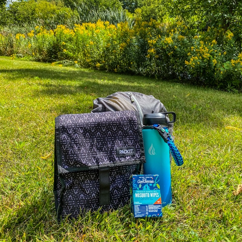 Backpack, lunch bag, mosquito repellent wipes and water bottle sitting in grass