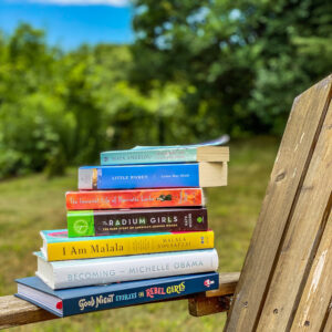 stack of books sitting on the arm of an Adirondack chair with green trees in background